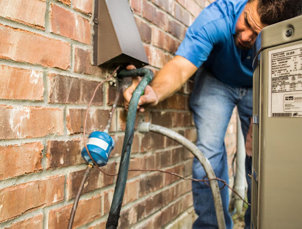 Technician repairing HVAC unit with focus on piping near brick wall.