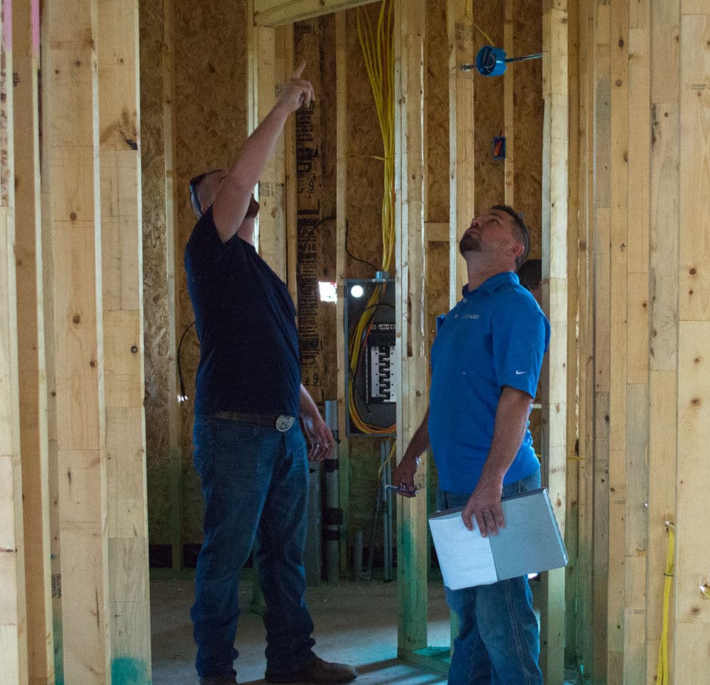 Two construction workers inspecting electrical wiring in a framed building interior.