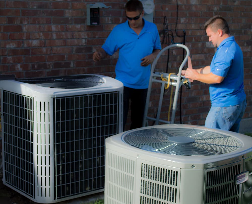 Two technicians servicing outdoor air conditioning units near a brick wall.